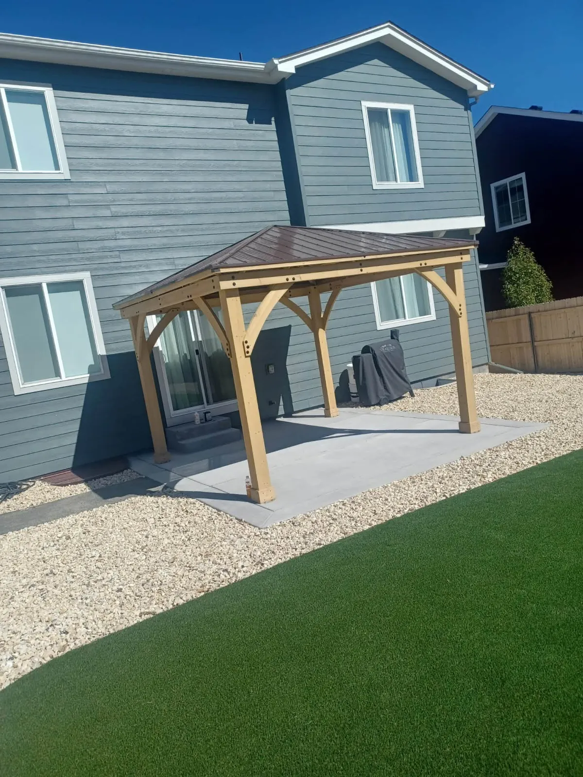 Timber gazebo over a concrete patio beside a gray house and turf lawn